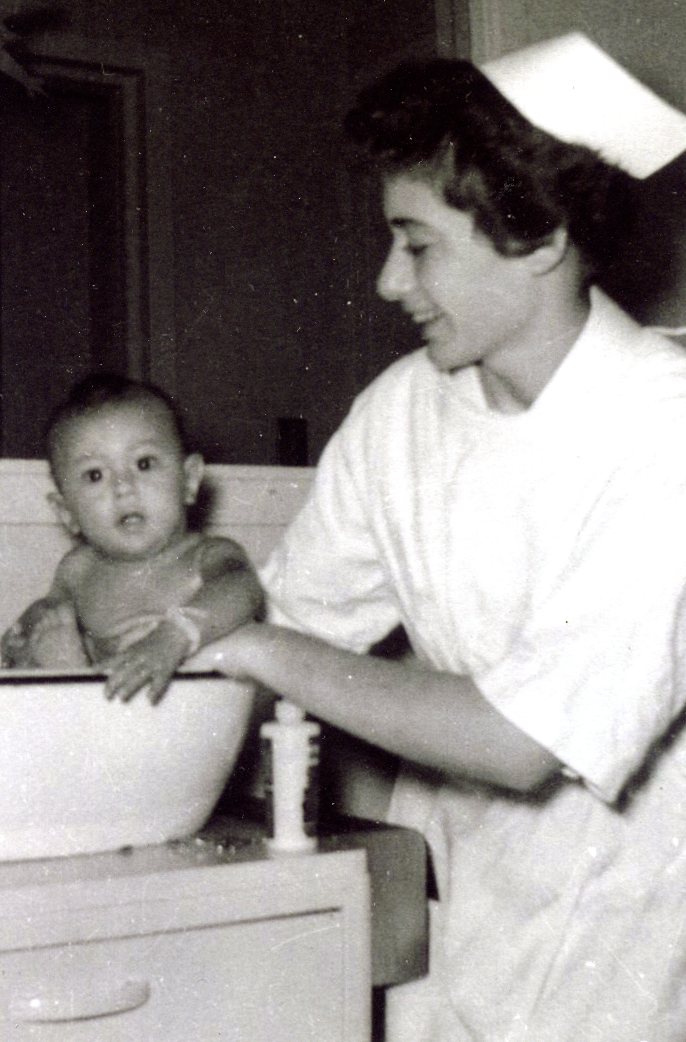 A smiling young woman in a nurses' uniform and cap bathes a baby in a basin.