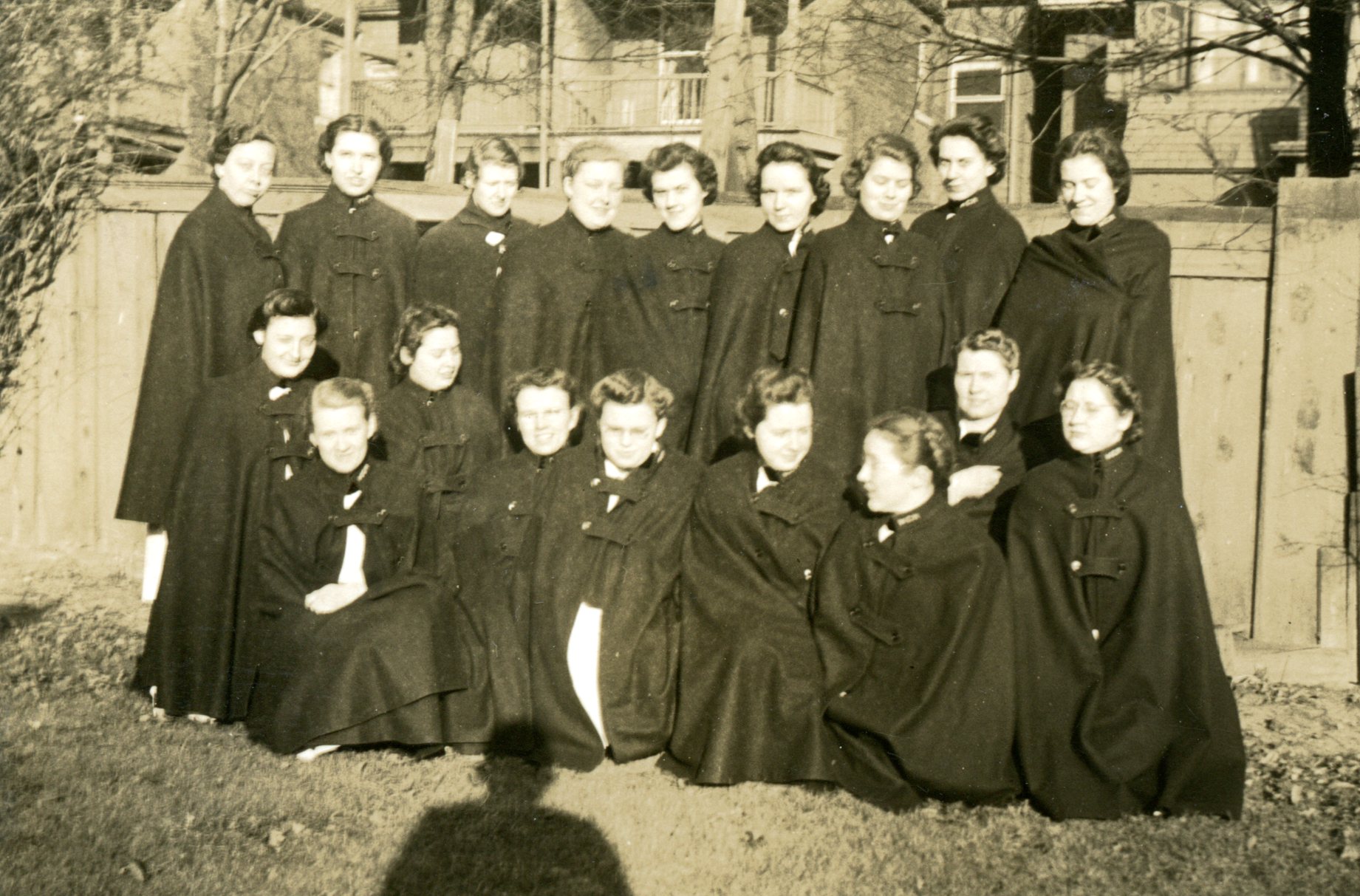 Eighteen young women pose for a portrait. They are wearing closed nurses' capes, and are outside before a fence. The photographer's shadow can be seen on the ground.