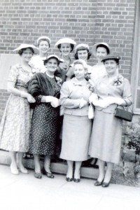 Eight women pose for a photo. They are dressed up, wearing long skirts, blouses, high heels, and hats. Flowers are pinned to their breasts.