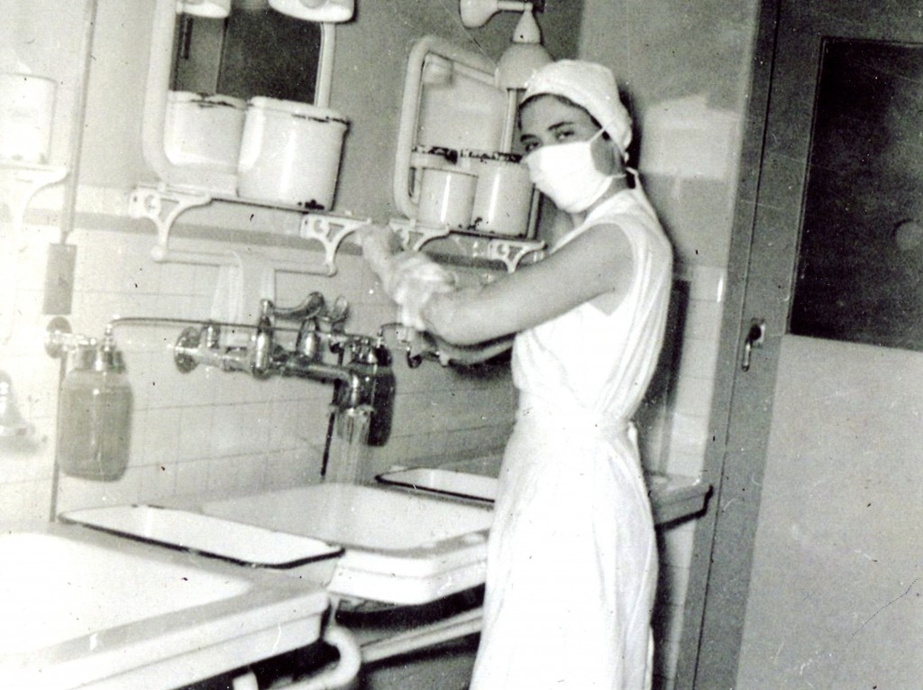 A young woman lathers up to her elbow at a sink. She is looking at the camera, and despite her surgical mask, her eyes are smiling. She is wearing elements of a nurses' uniform, though she is sleeveless. She wears her hair entirely covered in a protective cap.