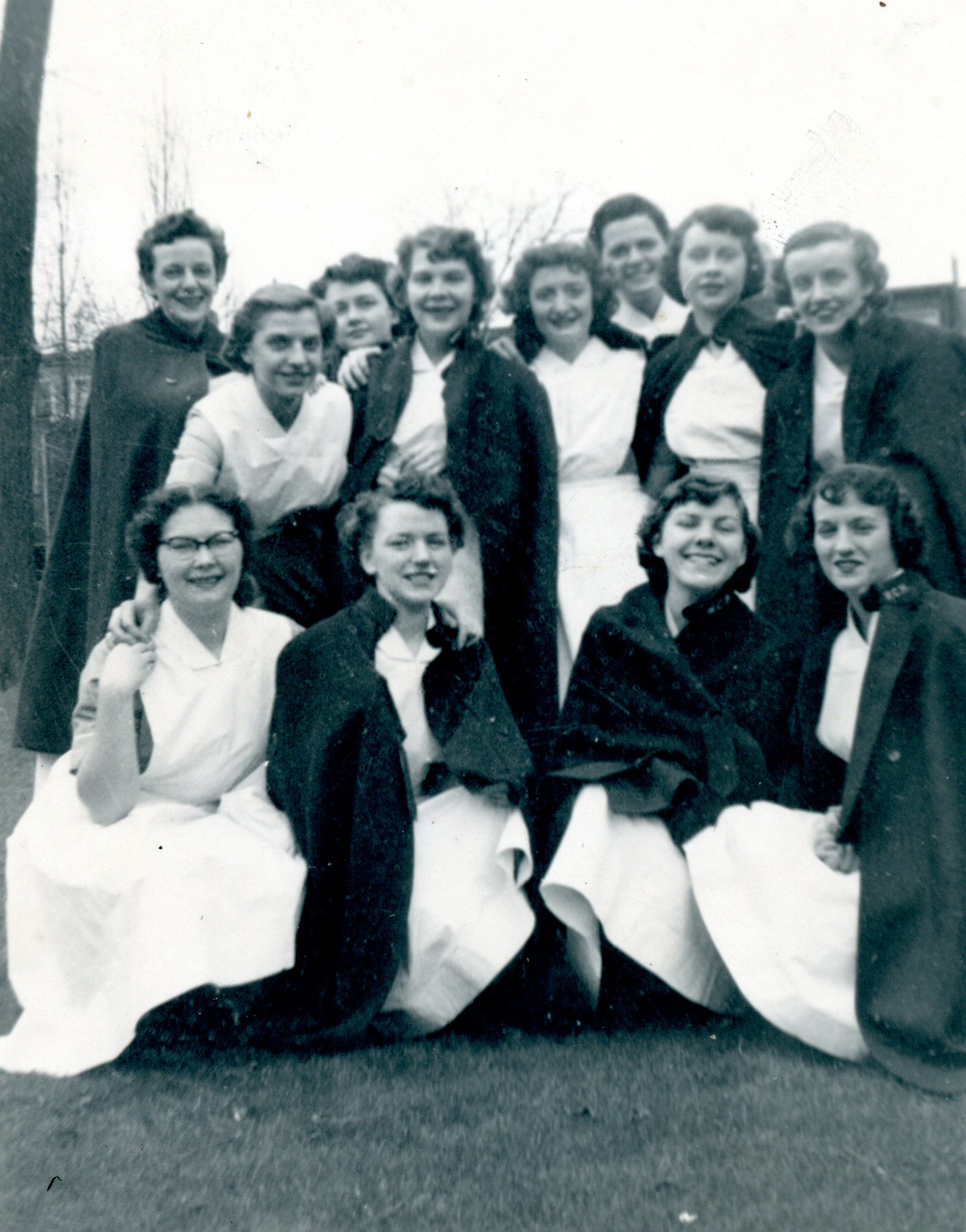 Twelve young women smile as they pose for the camera. They are outside, and most are wearing nurse's capes over their uniforms.