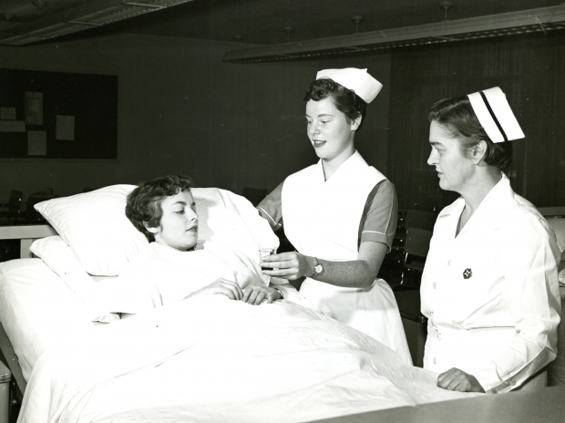 A young woman in a hospital gown lies in a bed, while another dressed in a nurse's uniform hands her a small glass. Another woman in a nurse's uniform with banded cap looks on.