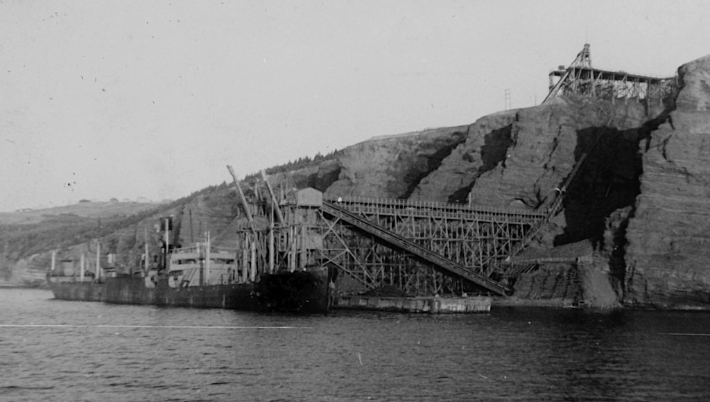 old photo of a cargo ship tied up at a loading pier with a conveyor belt