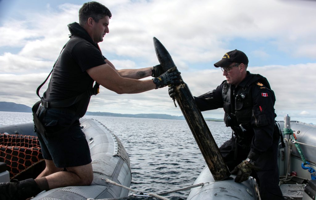 Two Navy divers lift an old artillery shell between two boats