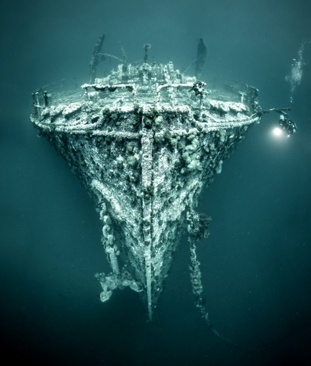 a scuba diver hovers underwater near the bow of a large sunken shipwreck