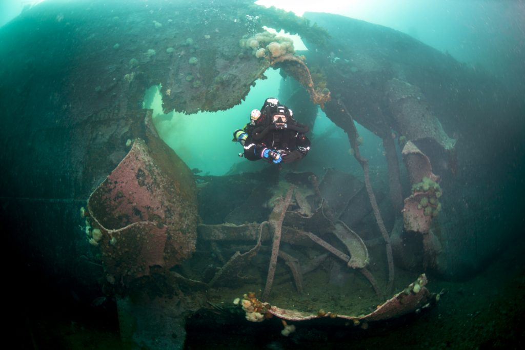 scuba diver swimming through a torpedo hole in sunken shipwreck