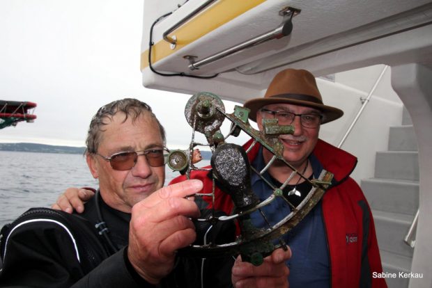 Diver holds a wet brass sextant while another man looks on smiling