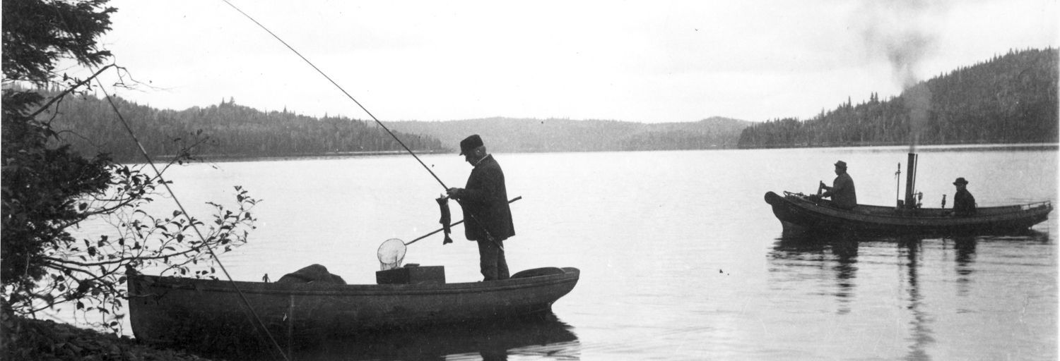 Black and white photo of fishermen in a steamboat on lac Édouard. In the foreground, a boat on the river’s edge; in it stands a man unhooking a fish from his rod.