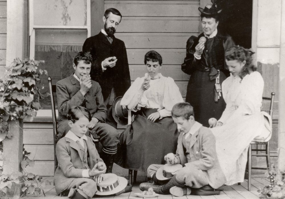 Black and white photo of a family sitting on a verandah. One man, two women, one girl, and three boys each looking at an apple in their hand.