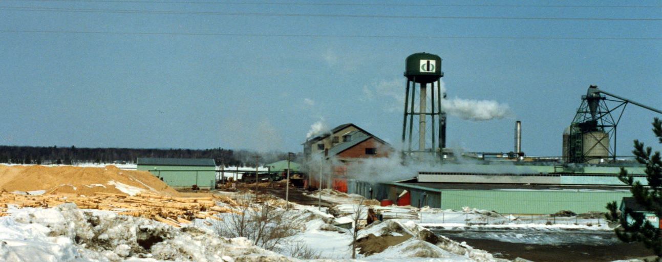 A brick saw mill building is surrounded by heaps of sawdust with a water tower and refuse burner in winter.