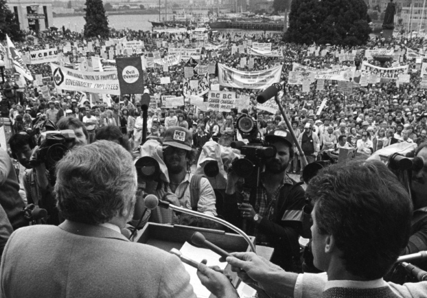 A man, speaking at a podium, faces out towards a large crowd of people on the lawn of the B.C. legislature. Reporters are gathered, holding microphones and cameras towards the podium. Organizational banners and protest signs are visible throughout the crowd.