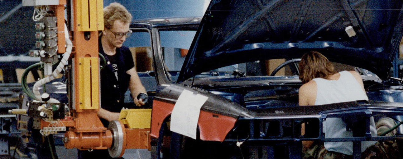 Colour image of three men using heavy machine tools to assemble a partially-completed car on an assembly line.