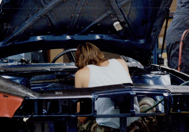 Colour image of three men using heavy machine tools to assemble a partially-completed car on an assembly line.