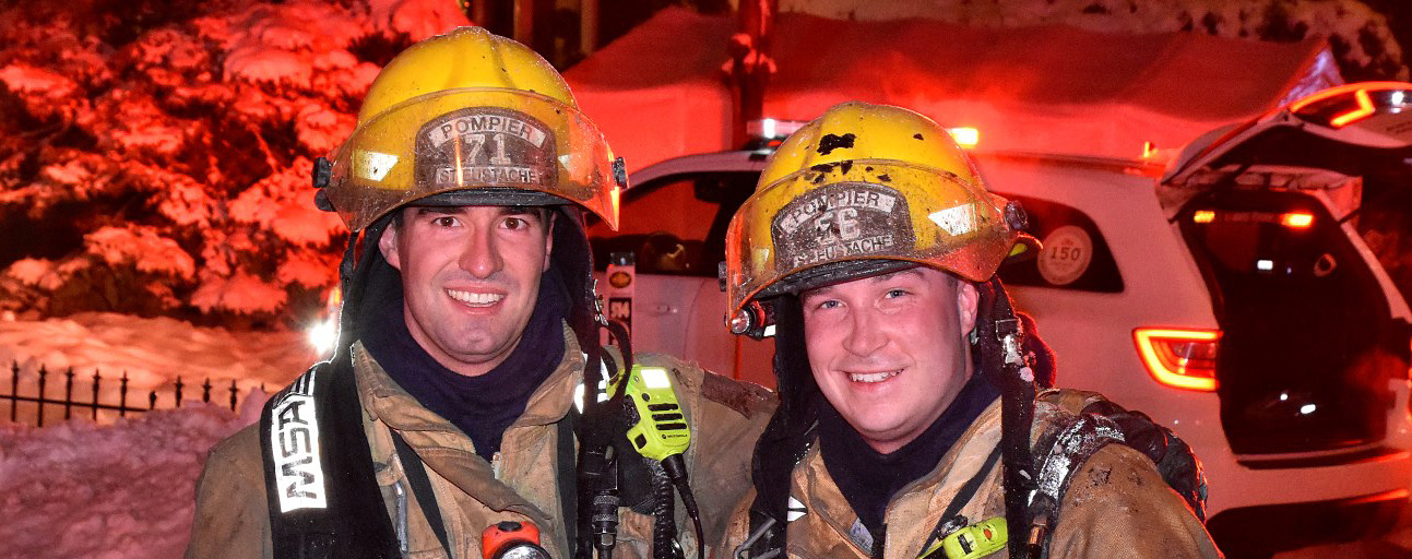 Photograph of two smiling firefighters in firefighting gear, in front of a car, a fire truck and many houses, at night, under the lights.