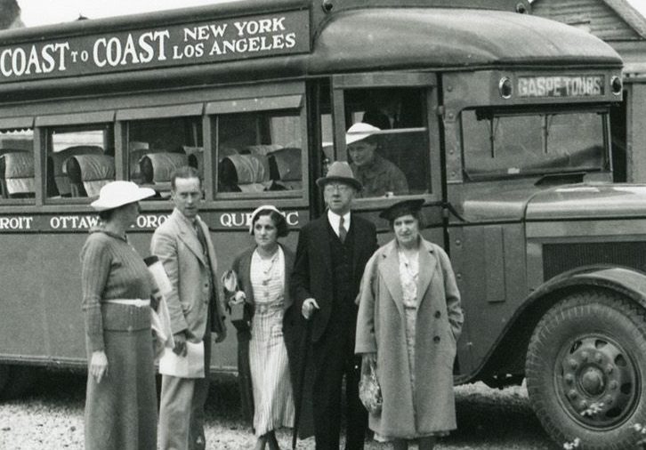 Black and white photograph of a group of people in front of a passenger bus in 1932. Behind the bus, you can see some houses in the village of Percé and the mountains that form the coast. On the bus is written Montreal Vancouver Coast to Coast New York Los Angeles.