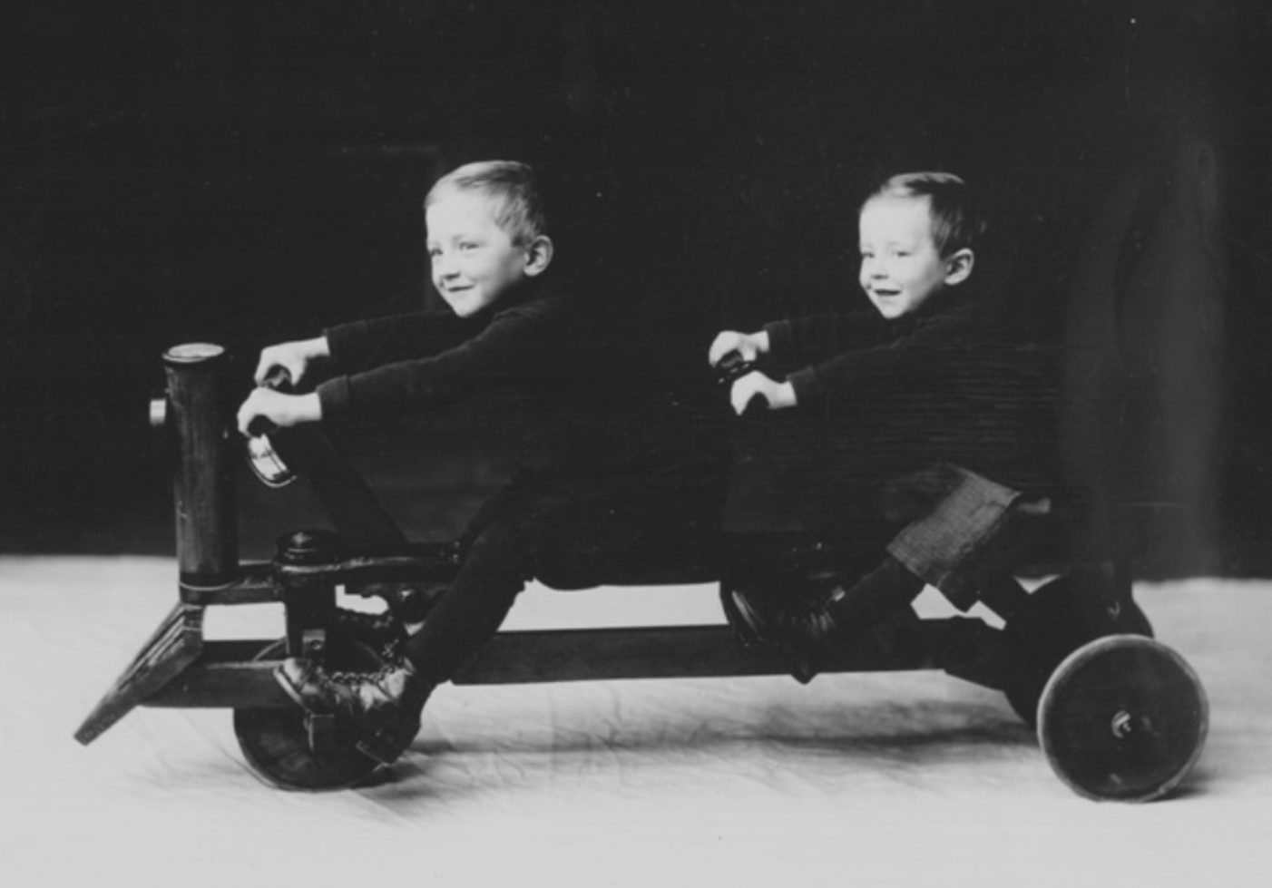 An old-fashioned black and white photograph of 2 smiling children seated on a cart that has 3 wheels and 2 sets of handlebars.