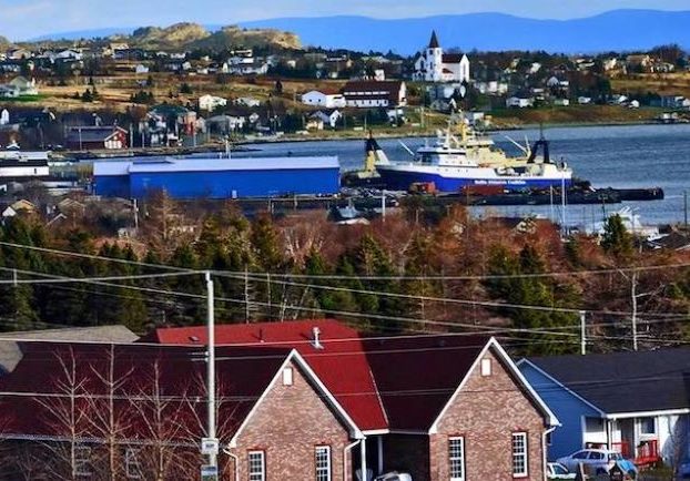 Colour photograph showing a ship in a bay with trees and homes wrapping around the bay in the foreground and background.