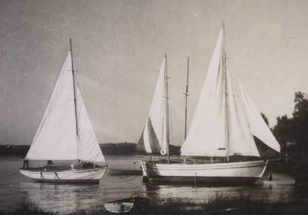 Black and white photograph of three white yachts, with hoisted sails, anchored near the shore.