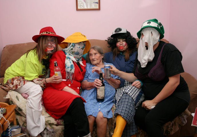 Four Mummers having a nip of rum sit with an elderly woman on a crowded couch