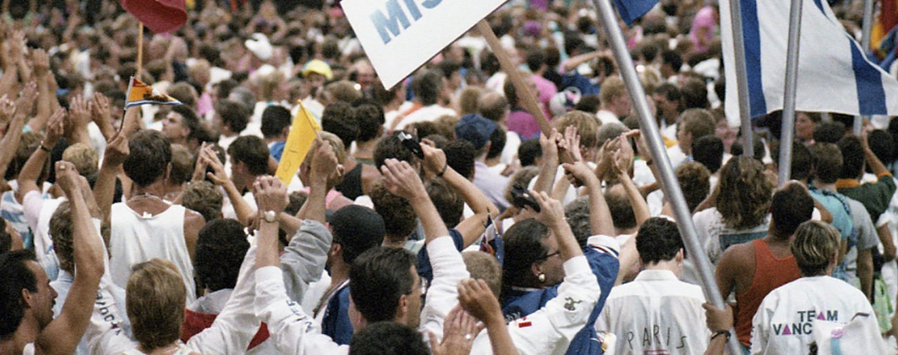 A large, tightly packed crowd of LGBTQ2S+ athletes and allies in B.C. Place Stadium, Vancouver, Canada, wave their arms and flags and sway to music at the Celebration ’90 Gay Games III Opening Ceremonies.