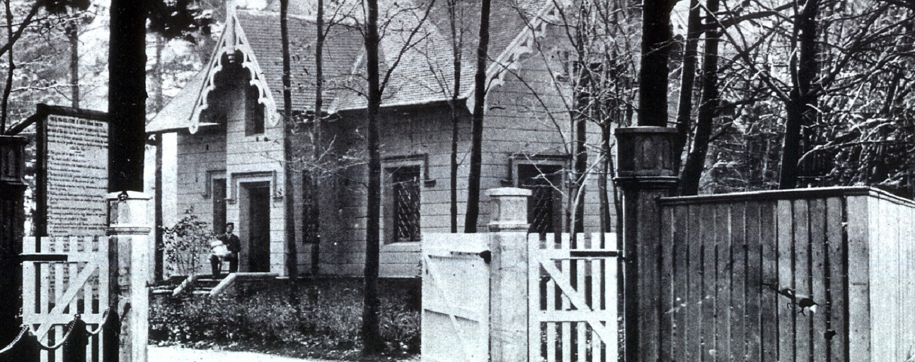 Black and white picture of a house surrounded by trees. A man is sitting on the front step holding a white-clothed baby. A large wood fence with open gates at the forefront.