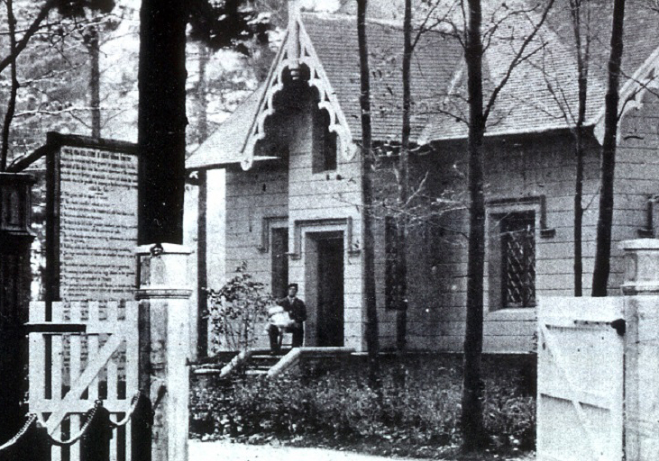 Black and white picture of a house surrounded by trees. A man is sitting on the front step holding a white-clothed baby. A large wood fence with open gates at the forefront.
