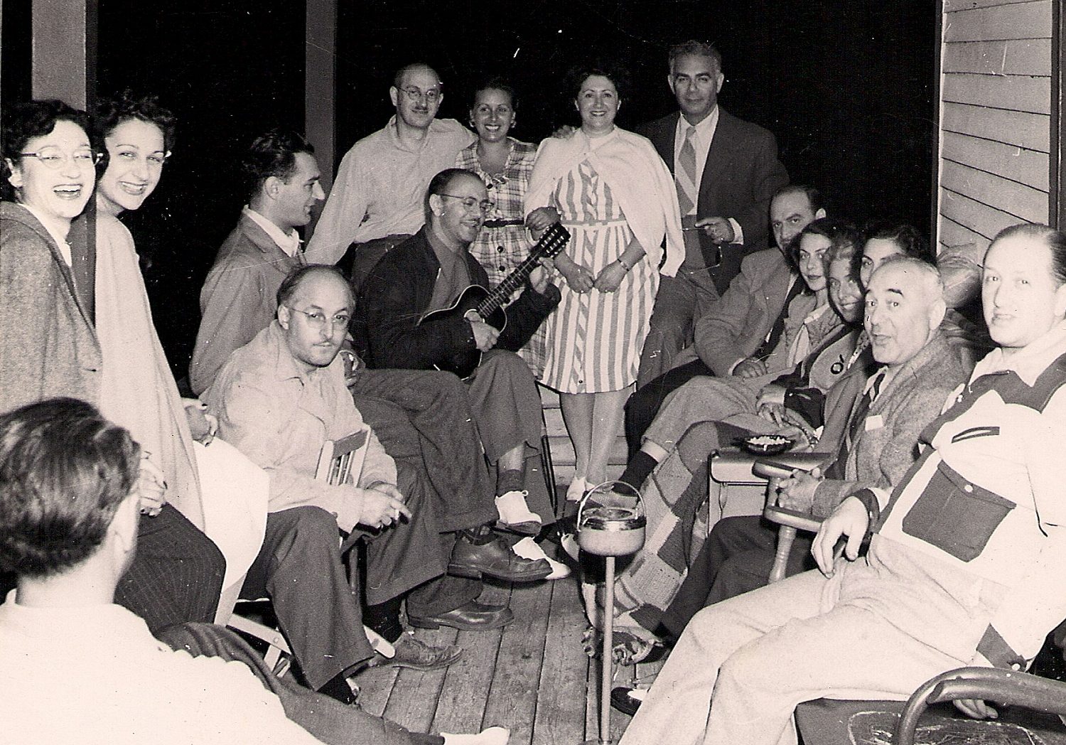 16 men and women seated on a wooden verandah – one man holding a ukelele