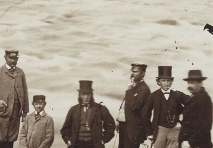 Black & white photograph of a group of nine people standing on the shore of a rushing river. At left, a man is perched partway up a tree trunk. Curé Labelle, a heavy-set man, is seen wearing a cassock and a wide-brimmed felt hat. At centre and right are six men and a teenaged boy wearing three-piece suits and top hats, caps or felt hats. Two of them are smoking pipes.
