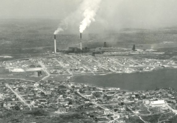 Black-and-white photograph of two cities build around a lake, separated by a vacant lot. In the background, you can see the smelter with three headframes and two smoking chimneys.