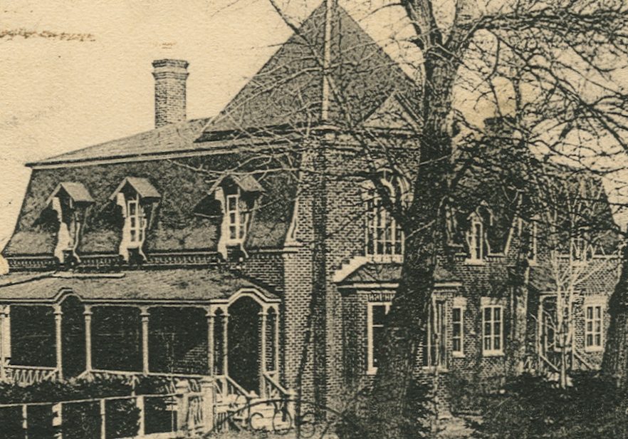 A black-and-white postcard showing a large elegant house with a veranda, bay window, and mansard roof. A few mature trees line the drive to the house.