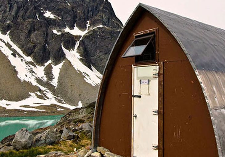 The Gothic Arch Hut front entrance with its brown front end-wall and bright white door sits next to the turquoise water of the glacial lake. A dark peak dotted with snow is visible in the background.