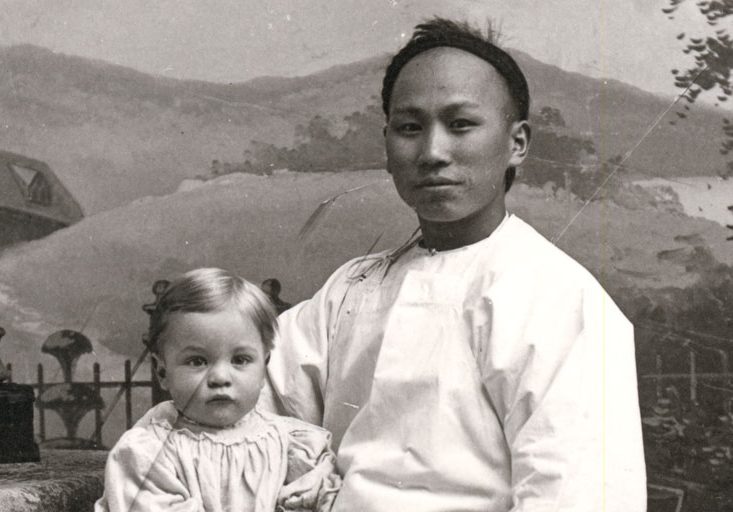 Chinese domestic worker sitting in a chair posing for a photograph with a young child on his right knee.