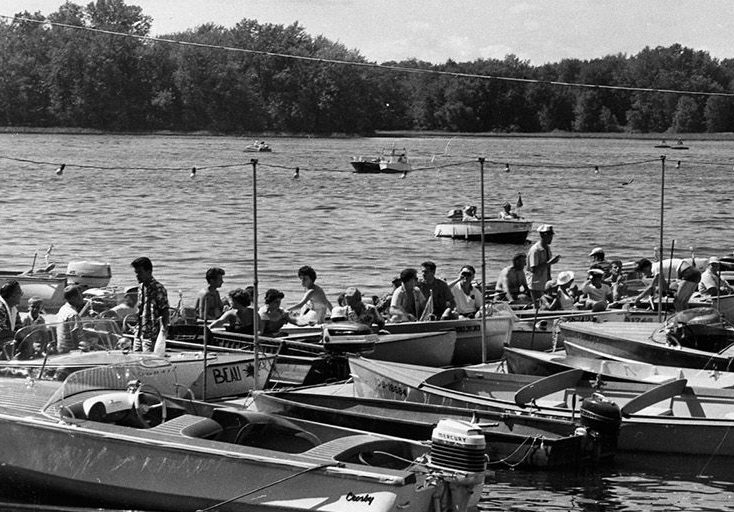Black and white photograph of small motorboats moored to a dock. Many people are sitting in them, ready for a boat parade.