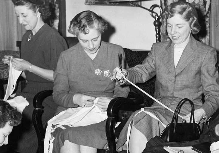 Women work on individual sewing projects in a black and white photo. Chairs have been added to the room to accommodate them. One woman sits on the floor in the left corner.