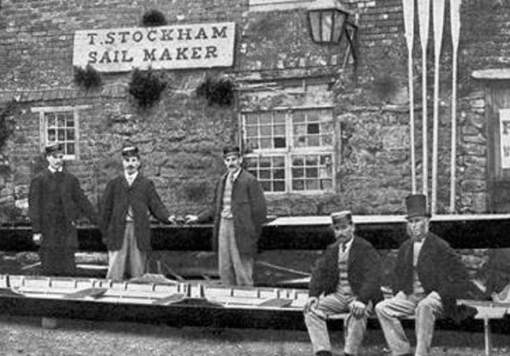 Image of the Paris Crew team members with their manager J. A. Harding posing in front of a boat house upon their arrival in Southampton, England. This black and white photo from July 1867 depicts a building made of stone blocks with their rowing scull positioned in front of the building, with rowing oars propped against its facade.