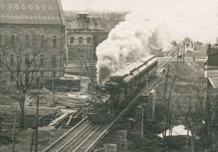 Photograph of a train travelling on a track with Almonte town hall in the background, 1950s