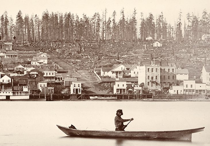 A sepia toned photograph of a man canoeing on the Fraser River in front of a recently deforested New Westminster. New Westminster has multiple buildings along the river, but the buildings become scarcer further up the hill. The upper portion of the hill is littered with tree stumps.