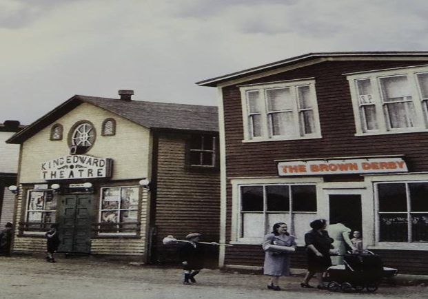 Colour archival photograph. Brown Derby, King Edward Theatre, A. Peckford, S. Cohen & Sons, and Purity Cafe. Kirk Pomeroy is carrying a birch broom, Daisy Bennett is wearing a blue-grey coat, and Mrs. Pomeroy is pushing a stroller.