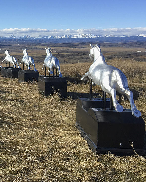 Colour photograph of landscape with eight silver coin-operated horses lined up in front of mountain panorama