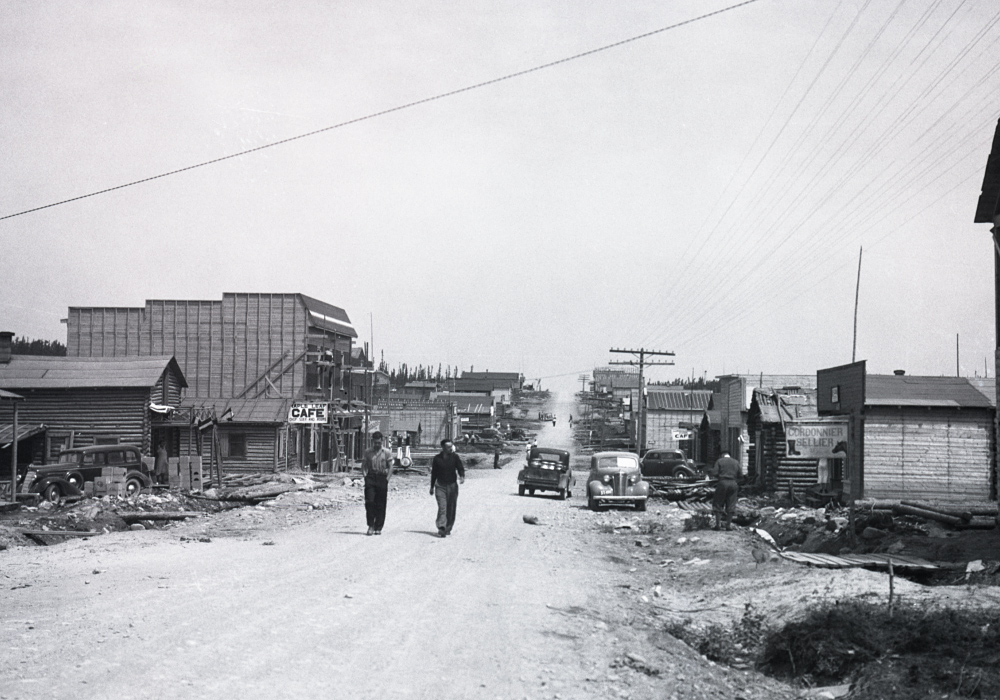 Black and white photograph of a gravel road lined with plank and log buildings. Two men walk toward the camera.