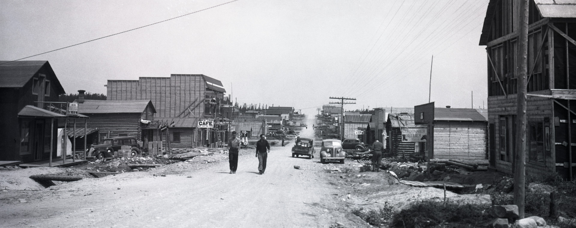 Black and white photograph of a gravel road lined with plank and log buildings. Two men walk toward the camera.