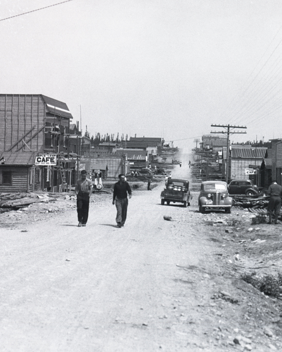 Black and white photograph of a gravel road lined with plank and log buildings. Two men walk toward the camera.