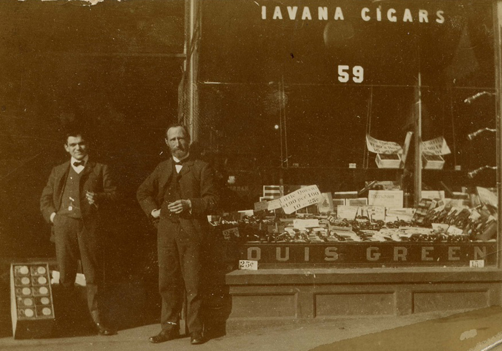 Storefront with large glass window with “Havana Cigars” written at the top of the window and “Louis Green” printed at the bottom – the window displays cigars and tobacco products – two men are standing in doorway.