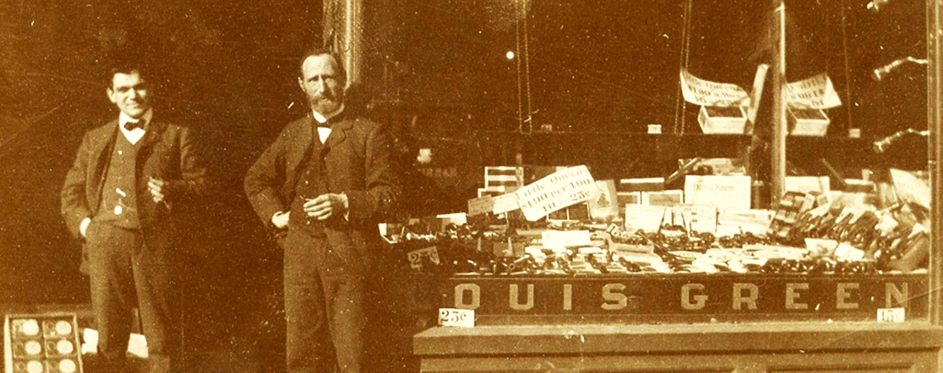 Storefront with large glass window with “Havana Cigars” written at the top of the window and “Louis Green” printed at the bottom – the window displays cigars and tobacco products – two men are standing in doorway.