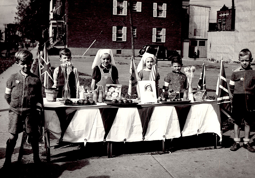 Portrait of six children, two girls in the middle and two boys on each side, behind a table with food, four Union Jacks and two vases of flowers.