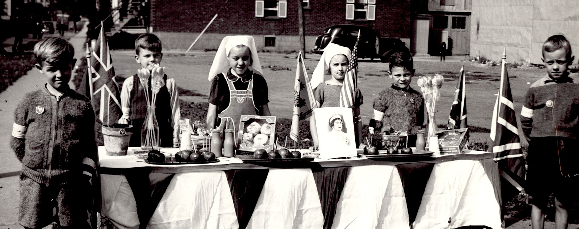 Portrait de six enfants, deux filles au centre et deux garçons de chaque côté, derrière une table remplie de nourriture, quatre drapeaux de l'Union Jack et deux vases de fleurs.