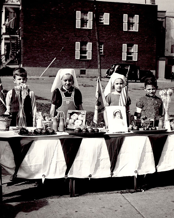 Portrait of four children, two girls in the middle and one boy on each side, behind a table with food, flags and two vases of flowers.