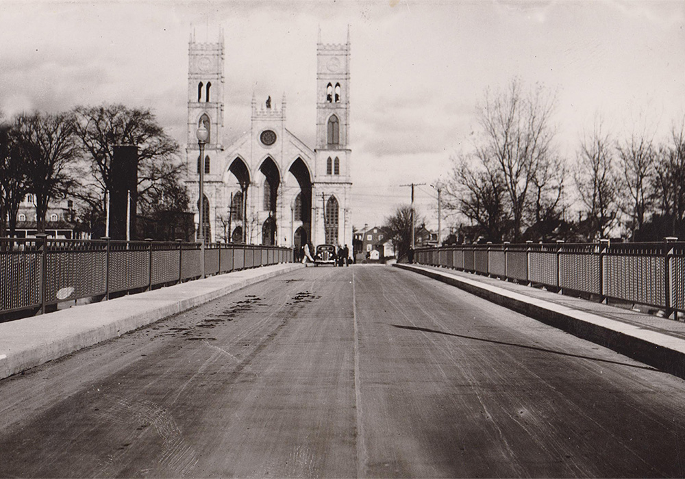 Black and white view of the span of a bridge at the end of which stands a church with two towers surrounded by old houses.