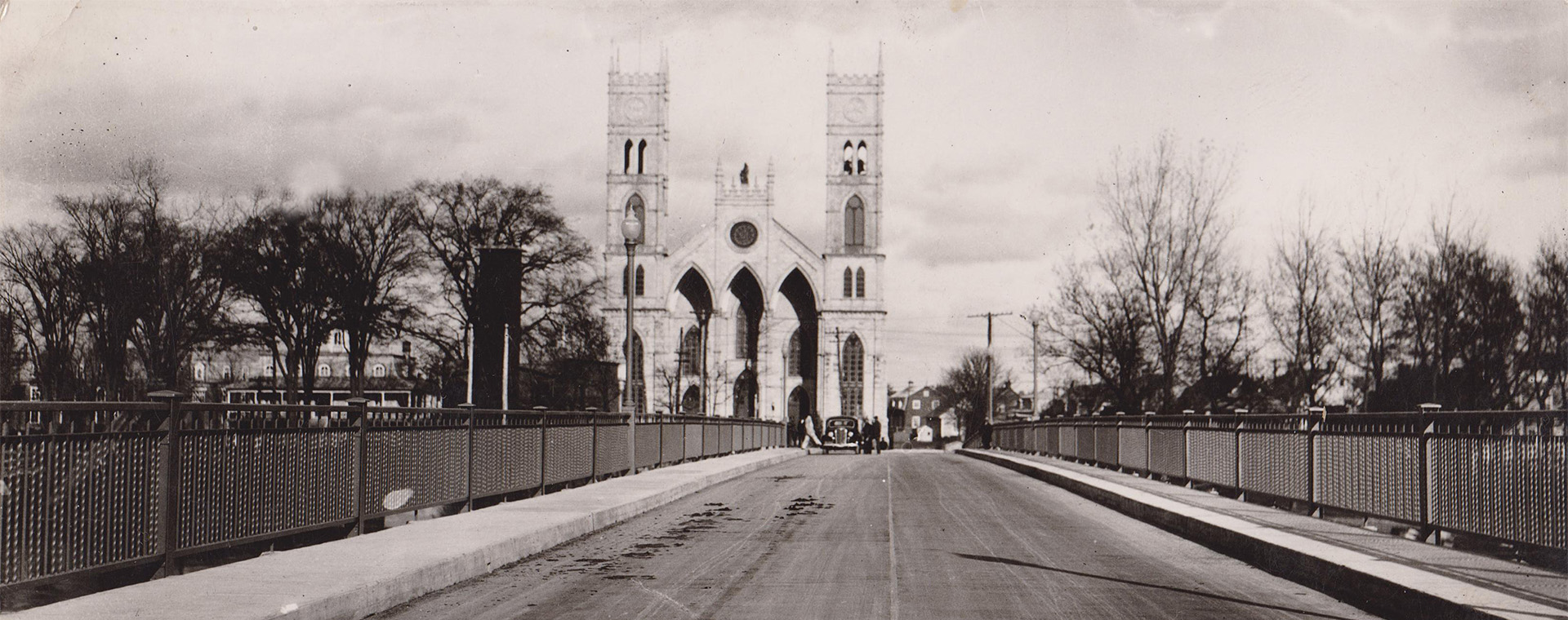 Black and white view of the span of a bridge at the end of which stands a church with two towers surrounded by old houses.