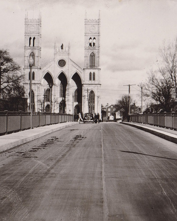 Black and white view of the span of a bridge at the end of which stands a church with two towers surrounded by old houses.